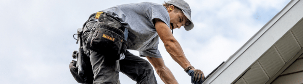 A man with a tool belt on a roof with a pencil in his mouth while measuring a shingle
