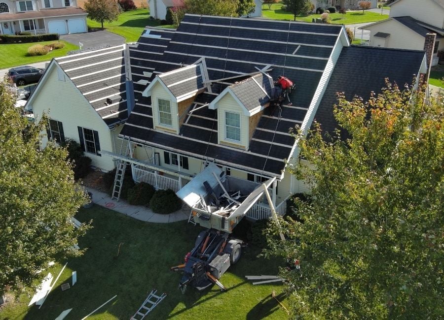 Aerial view of roofers replacing a house roof, with ladders and a debris container below